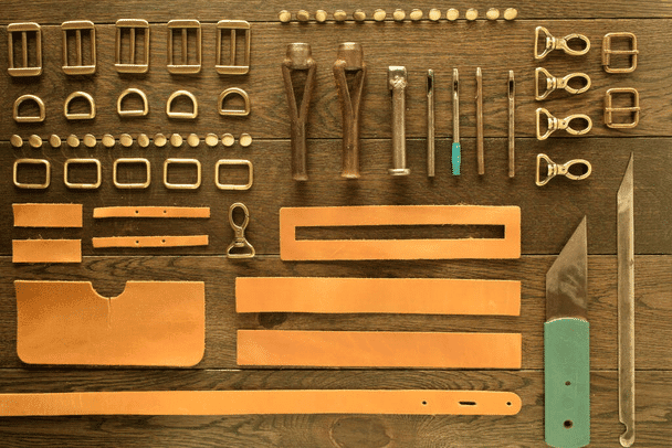 Assorted leathercraft tools and accessories, including buckles, buttons, leather strips, and knives, are neatly arranged on a wooden surface.