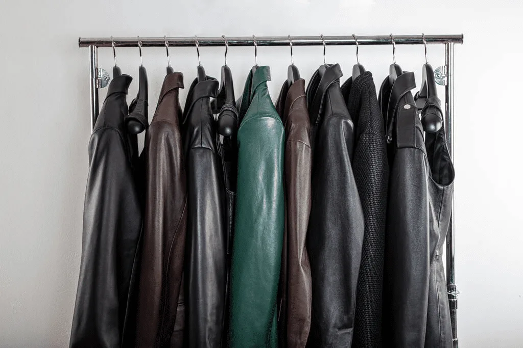 A row of leather jackets, mostly black and brown with one green jacket, showcase expert leathercraft as they hang on a metal clothing rack, perfectly aligned for optimal leather storage.