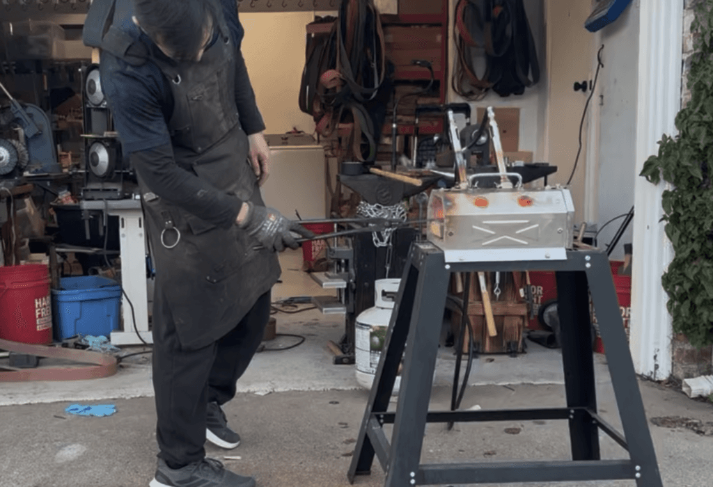 A person in an apron skillfully forges metal using a furnace in an outdoor workshop, where various tools and equipment are visible in the background.