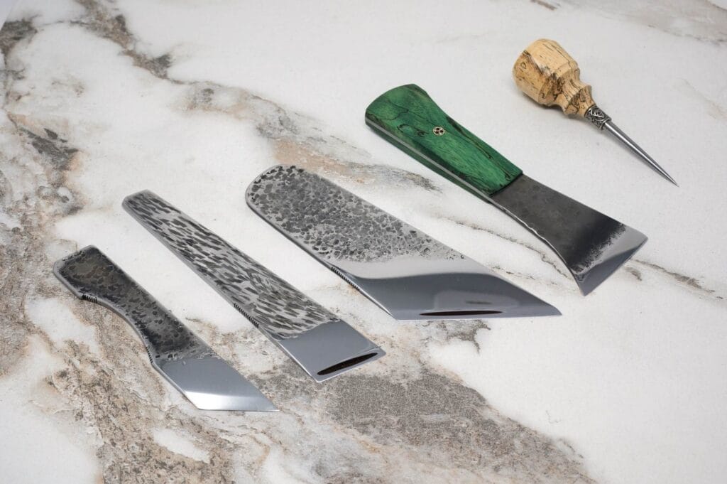 Four uniquely shaped knife blades and a decorative awl with a green handle are displayed on a marbled surface.