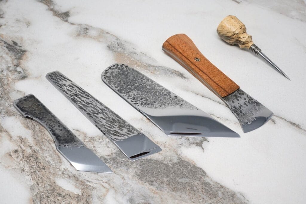 Four artisan knives with textured blades and wooden handles are displayed on a marble surface.