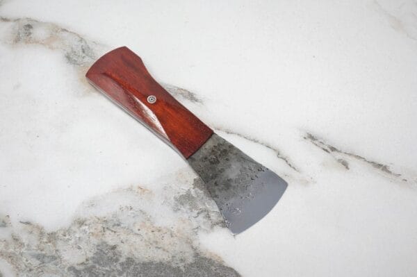 A small hatchet with a wooden handle and metal blade, reminiscent of the Flat American Signature Leathercraft Knife, rests on a snowy surface.