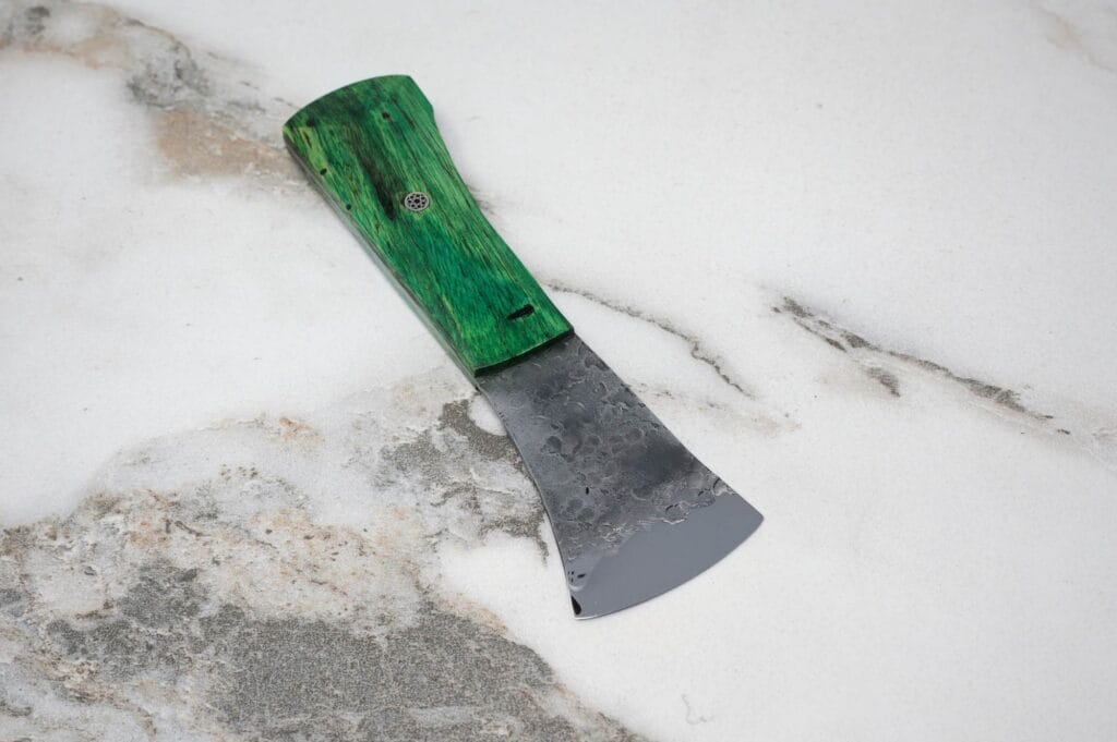 The Forged American Signature Classic, a small axe with a green handle and weathered blade, rests on a textured stone surface.