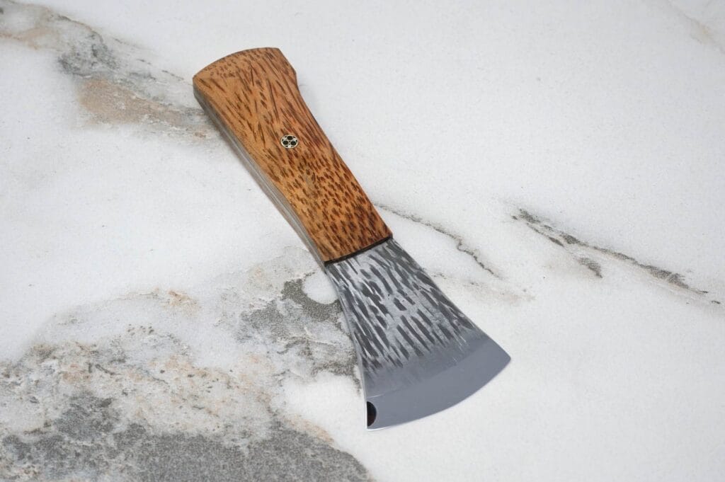 A small hand axe with a wooden handle and textured metal blade, reminiscent of the Forged American Signature Classic, rests on a marble surface.