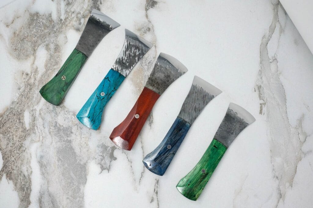 Five colorful handled scrapers with metal blades are arranged on a marbled surface.