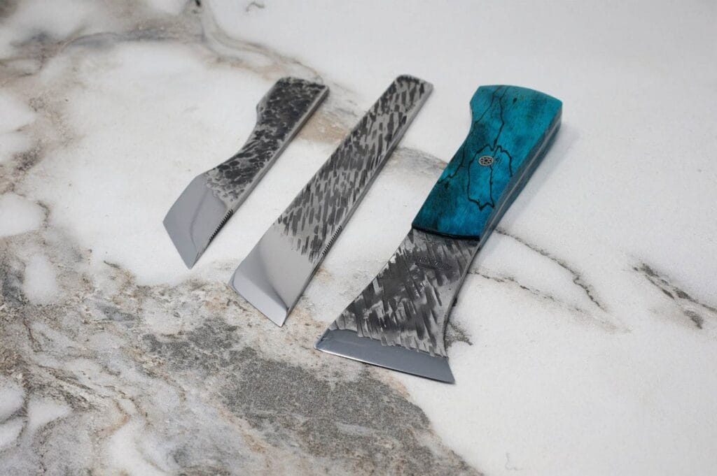 Three chisels on a marble surface, differing in size and shape. One has a turquoise handle, while the others are entirely metal.