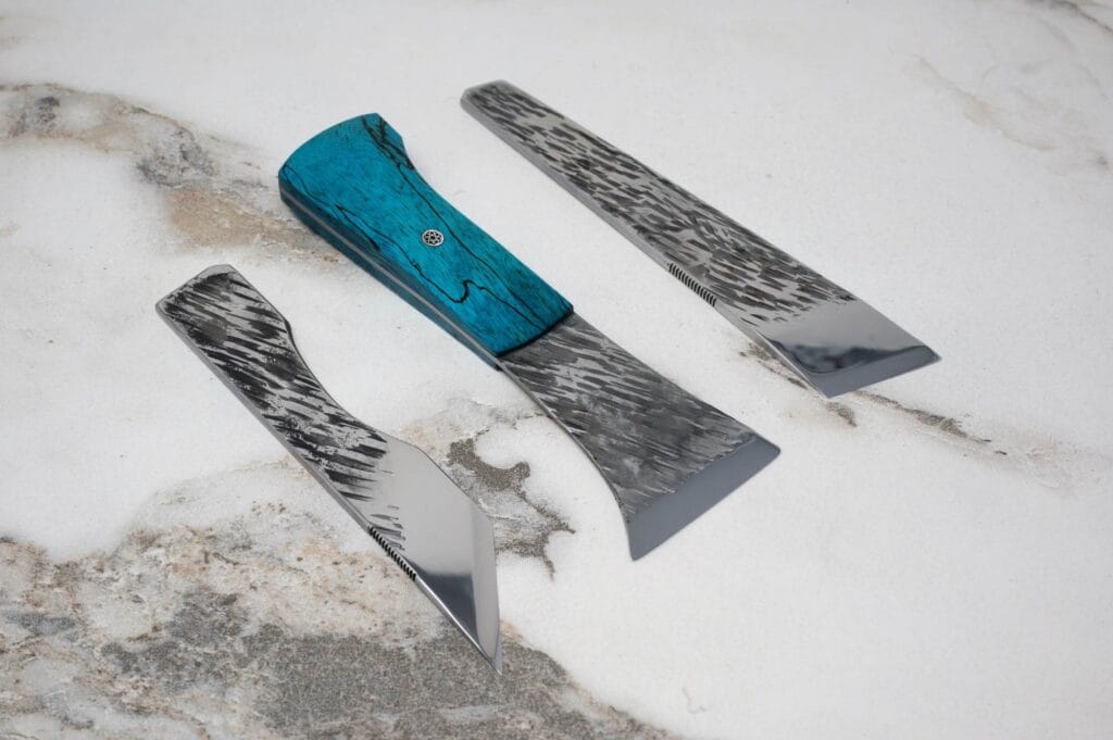 Three metal blades with patterned designs are arranged on a textured surface. The blade handles are blue, contrasting with the silver and patterned knife edges.