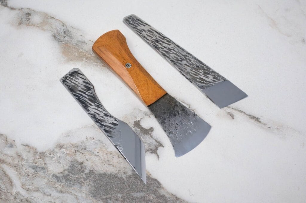 Three knives with patterned metal blades and wooden handles are laid out on a textured marble surface.