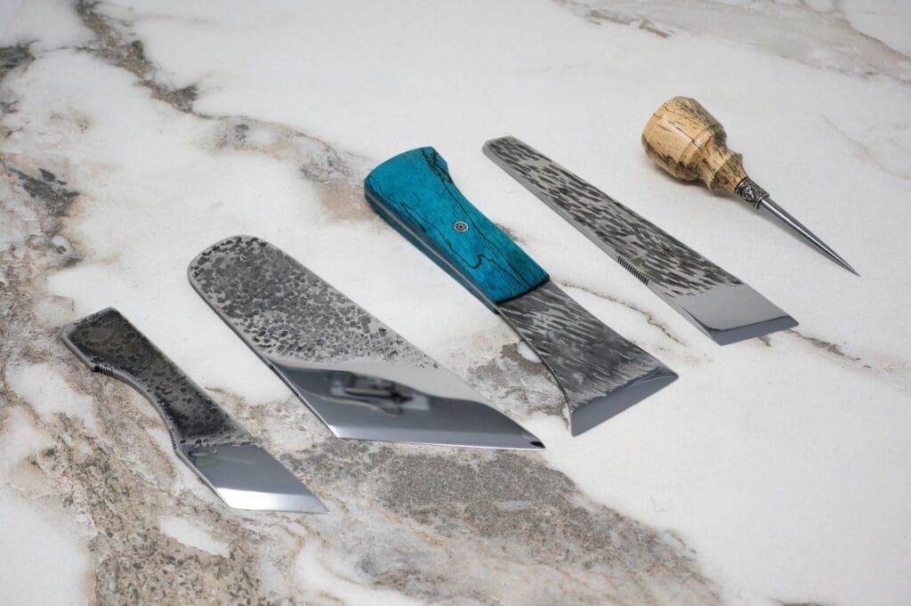 A set of knives with textured metal blades and colorful handles are arranged on a marbled surface.