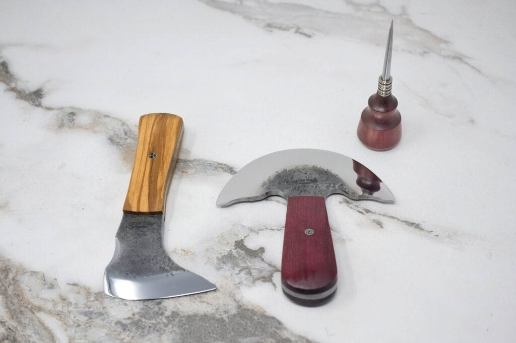 A hatchet, a curved knife with a wooden handle, and a small awl with a wooden grip are laid on a marble surface.
