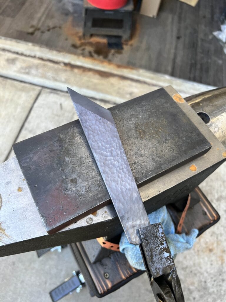 A knife blade on an anvil with a textured surface, accompanied by a blue cloth, creates a vivid description of tools and craftsmanship.