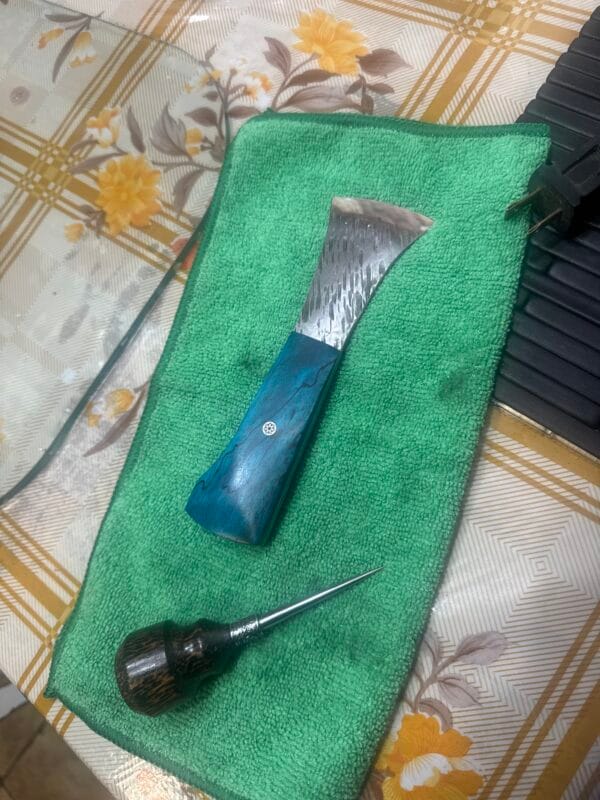 A Signature Addon - Mercer chisel with a blue handle and a wood-handled awl rest on green cloth atop a patterned table—an excellent addition to any craftsman’s toolkit.
