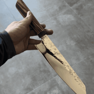 A hand holds a large, reflective kitchen knife with a wooden handle over a gray tiled floor, showcasing the results of our Kitchen Knife Sharpening Services.